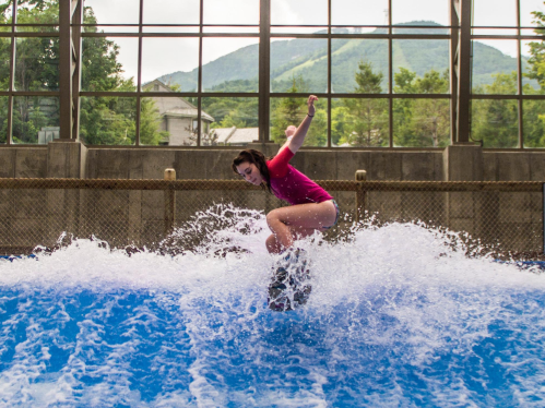 A person surfing on a wave in an indoor facility, with mountains visible through large windows in the background.
