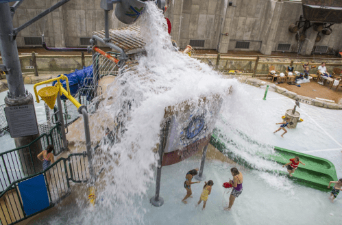 Children play in a water park area as a large splash of water cascades down from a play structure.