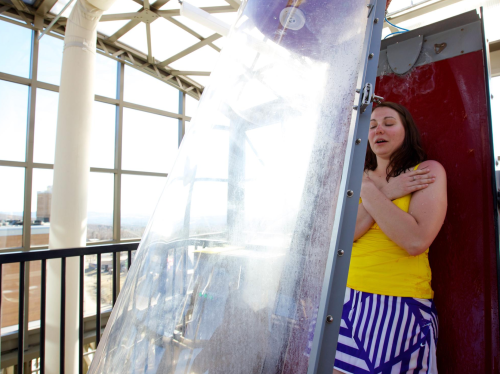 A woman in a yellow top stands inside a transparent water slide structure, looking excited as water cascades around her.