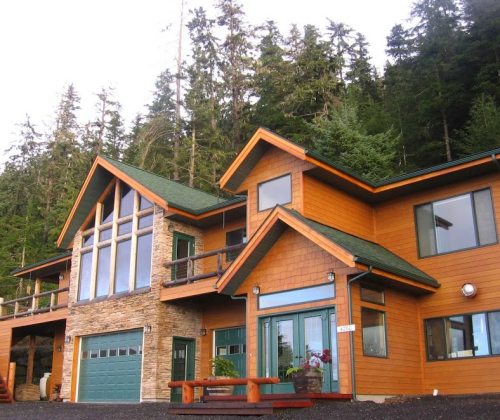 A modern wooden house with large windows, surrounded by tall trees and a gravel driveway.
