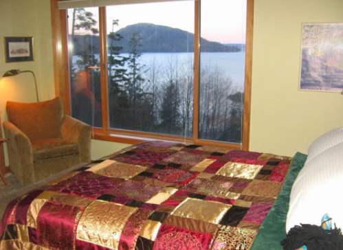 Cozy bedroom with a patchwork quilt, armchair, and large window showcasing a scenic lake view and mountains.