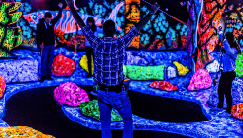 A group of people playing mini-golf in a vibrant, neon-lit environment with colorful decorations.