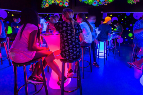 A vibrant, neon-lit room with children sitting at glowing tables, enjoying drinks and socializing.