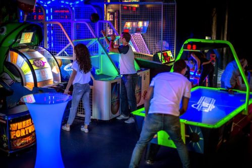 A vibrant arcade scene with people playing games under colorful lights, including a spinning wheel and air hockey.