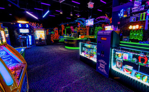 A vibrant arcade with neon decor, featuring games, ticket counters, and colorful prizes under blacklight.