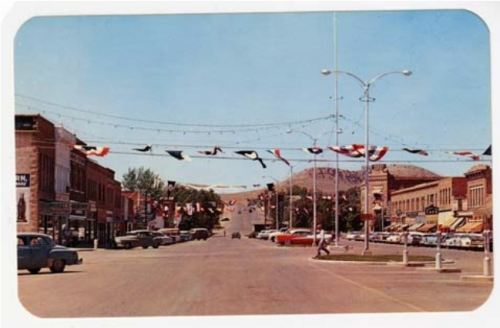 A vintage street scene with decorated buildings, flags, and classic cars under a clear blue sky.