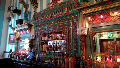 Colorful bar with ornate decorations, shelves filled with various bottles, and vibrant lighting.
