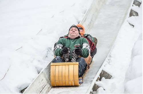 Two people sledding down a snowy track, one in front with a focused expression and the other behind, enjoying the ride.