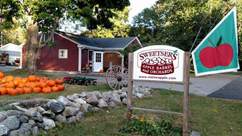 A red barn with pumpkins in front, featuring a sign for Sweetser's Apple Barrel and Orchards and an apple-themed flag.