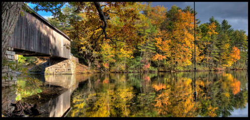 A scenic view of a covered bridge over a calm river, surrounded by vibrant autumn foliage reflecting in the water.