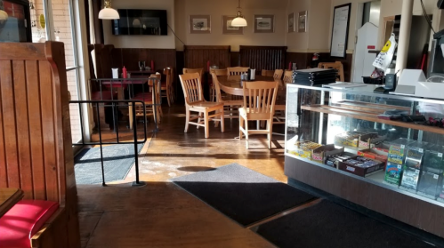 Interior of a restaurant with wooden tables and chairs, a display case, and sunlight streaming through the windows.