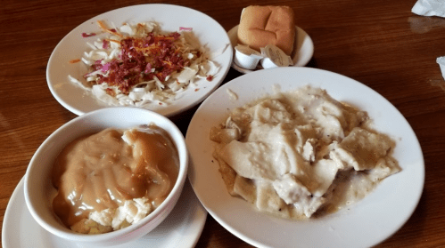 A plate with chicken and dumplings in gravy, a side of coleslaw, mashed potatoes with gravy, and a dinner roll.