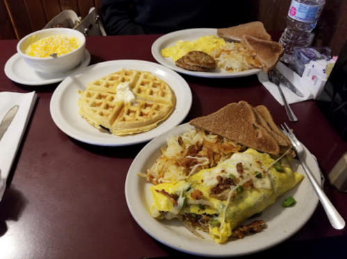 A table with a plate of omelette, hash browns, toast, a waffle, and a bowl of yellow cereal.