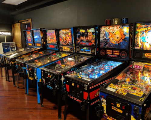 A row of colorful pinball machines in a dimly lit arcade, showcasing various game themes and designs.