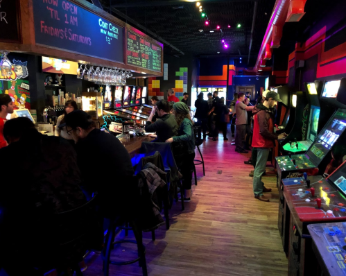 A lively arcade bar with people enjoying games, drinks, and a vibrant atmosphere filled with colorful lights.