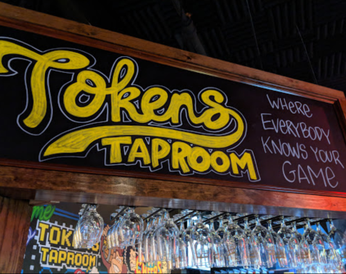 Sign for "Tokens Taproom" with the tagline "Where Everybody Knows Your Game," above a row of hanging glasses.