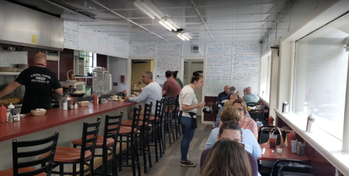 A busy diner with patrons seated at a long counter and tables, while staff prepare food in the kitchen. Walls covered in menus.