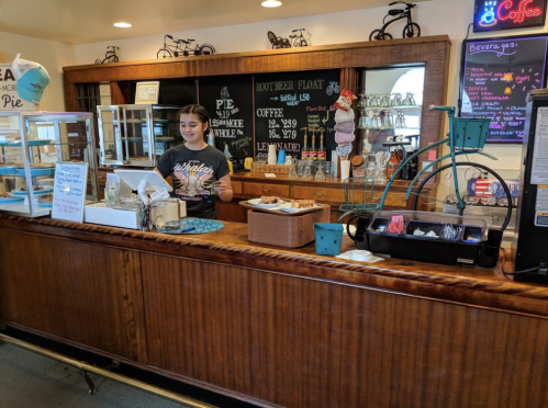 A young woman stands behind a wooden counter in a cozy café, surrounded by desserts and a menu board.