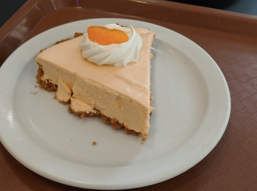A slice of peach mousse cake on a white plate, topped with whipped cream and a slice of peach, on a brown tray.