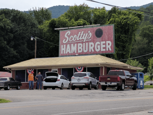 A roadside hamburger restaurant with a large sign reading "Scotty's Hamburger," featuring parked cars and people outside.
