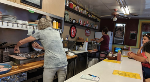 A diner scene with two women cooking at the grill, while customers sit at the counter. Condiments are visible on the table.