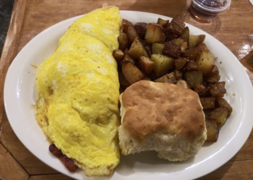 A plate with a fluffy omelet, crispy potatoes, and a biscuit, served with a small cup of syrup.