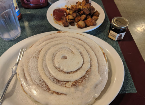 A large cinnamon roll pancake with icing, served with a side of diced potatoes and a small jar of syrup.