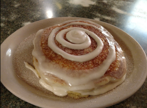 A freshly baked cinnamon roll drizzled with white icing on a plate, sprinkled with sugar.