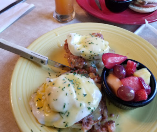 A plate of eggs Benedict with hollandaise sauce, served with a side of mixed fruit in a small bowl.