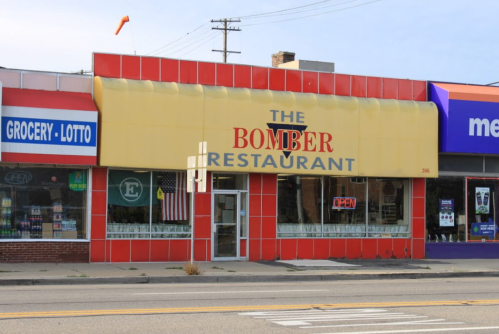 A storefront of "The Bomber Restaurant" with a yellow awning and large windows, located next to a grocery store.