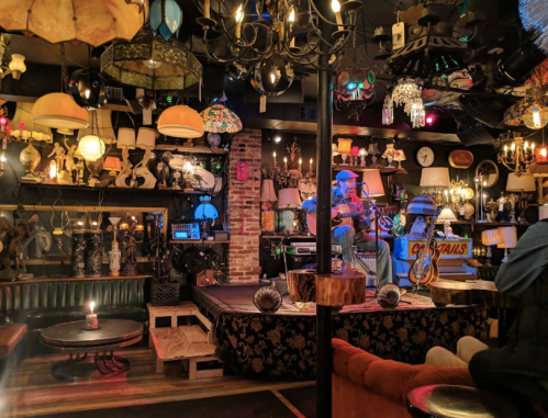 A cozy bar with eclectic decor, featuring a musician performing on stage surrounded by various lamps and vintage items.