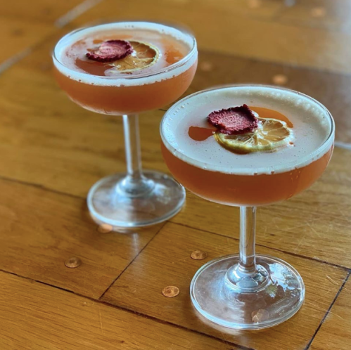 Two elegant cocktails in coupe glasses, garnished with dried rose petals and lemon slices, on a wooden table.
