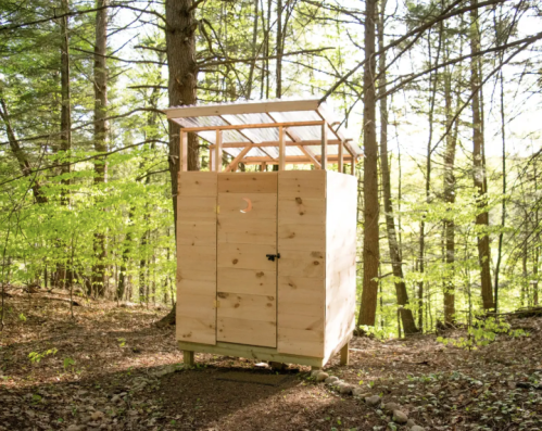 A wooden outdoor shed with a slanted roof, surrounded by trees in a sunlit forest.