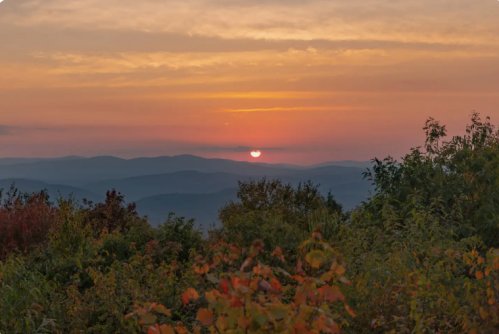 A sunset over rolling mountains, with vibrant autumn foliage in the foreground and a colorful sky.