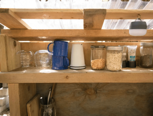 A wooden shelf displaying various jars, mugs, and a small white container in a rustic kitchen setting.