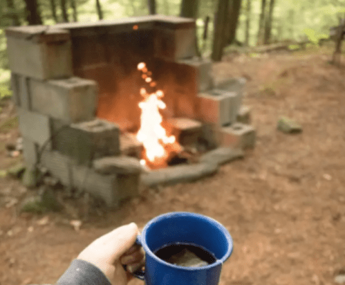 A hand holding a blue mug in front of a stone fire pit with flames, surrounded by trees and forest floor.