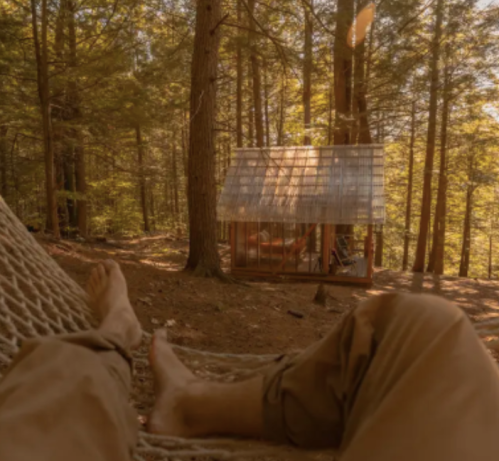 A person relaxes in a hammock with bare feet, surrounded by trees and a cozy cabin in the background.