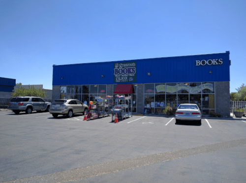 A blue storefront labeled "Grassroots Books" with parked cars and outdoor book displays on a sunny day.