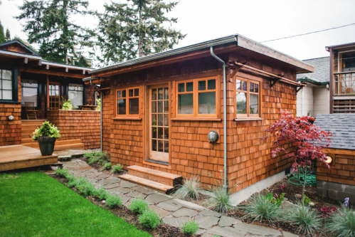 A cozy wooden cabin with a shingled exterior, surrounded by greenery and a stone pathway.