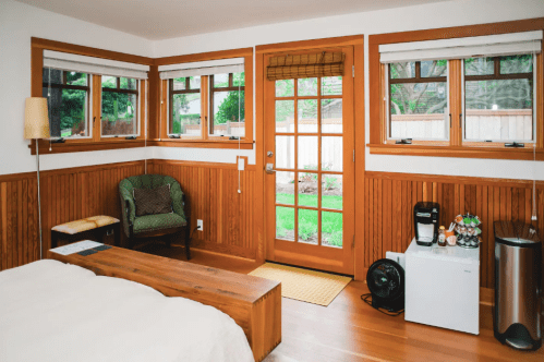 Cozy bedroom with wooden accents, large windows, a bed, a small fridge, and a coffee maker near a glass door to the garden.