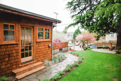 A cozy wooden house with a stone pathway, surrounded by green grass and trees, leading to a colorful garden.
