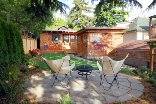 A cozy backyard with two chairs around a fire pit, surrounded by greenery and a wooden cabin in the background.