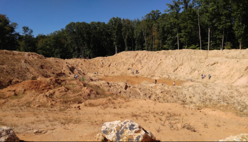 A large, sandy excavation site with people working in the background, surrounded by trees and blue sky.