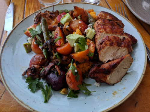 A plate of grilled chicken with a colorful salad of roasted vegetables, avocado, and fresh herbs.