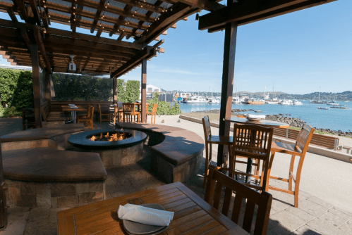 Outdoor dining area with wooden tables, a fire pit, and a view of boats in the water under a clear blue sky.