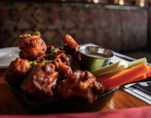 A bowl of crispy chicken wings garnished with green onions, served with celery sticks and a small cup of dipping sauce.