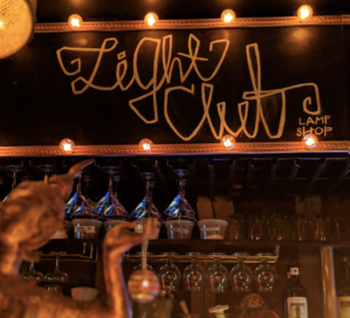 A dimly lit bar with a sign reading "Light Club" and shelves of glassware and lamps in the background.