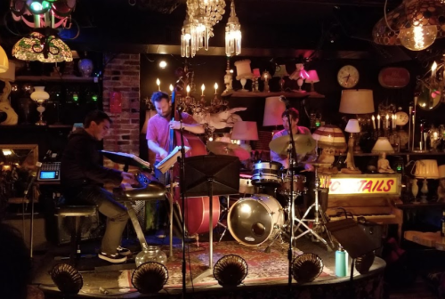 A jazz trio performs on stage in a cozy, dimly lit venue filled with eclectic decor and vintage lamps.