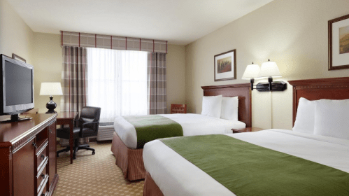 A cozy hotel room with two beds, a TV, a chair, and natural light from a window with plaid curtains.
