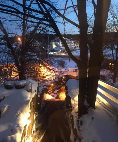 A snowy landscape at dusk, featuring trees, a stream, and softly lit buildings in the background.